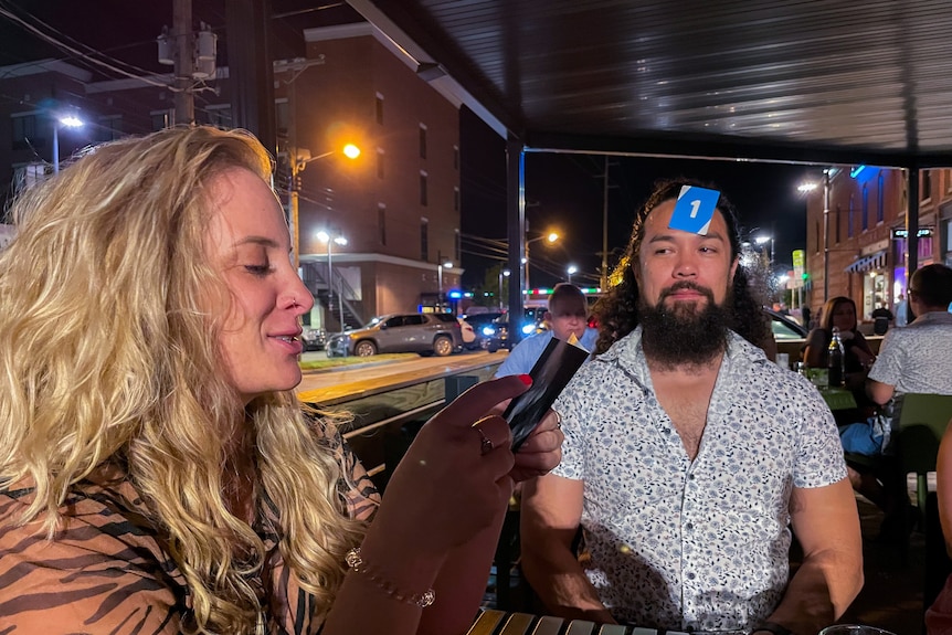 A man sits in a beer garden with a Uno card stuck to his forehead 