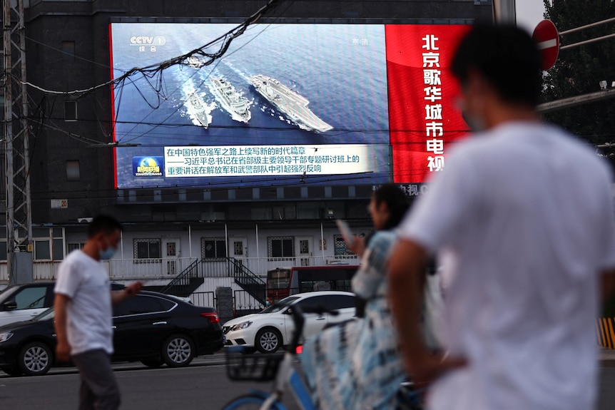 People in the street look up at a screen showing a news bulletin with images of Chinese warships