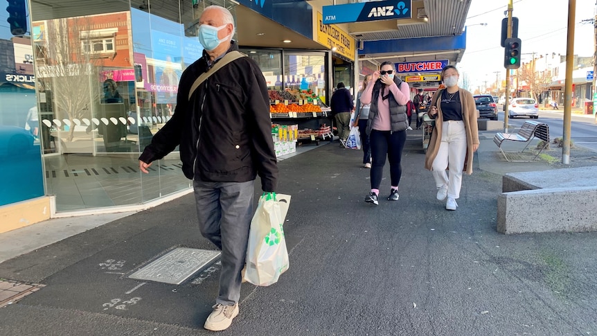 A man wearing a mask walking along a shopping strip with a plastic bag in hand. 