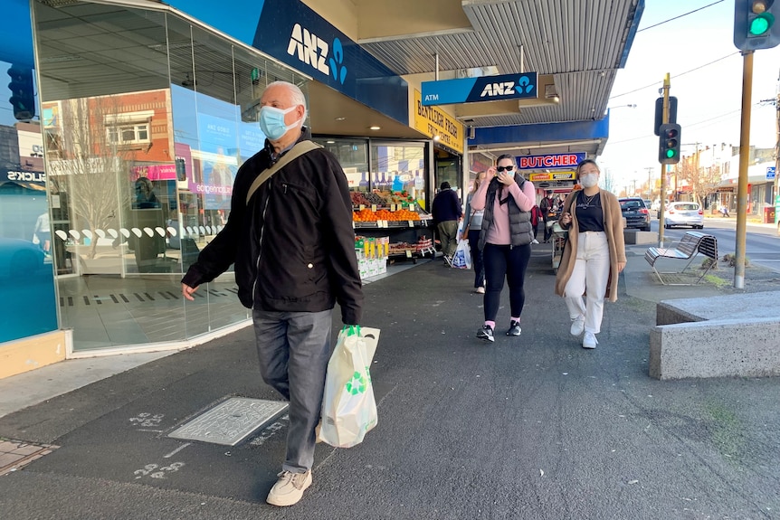 A man wearing a mask walking along a shopping strip with a plastic bag in hand. 