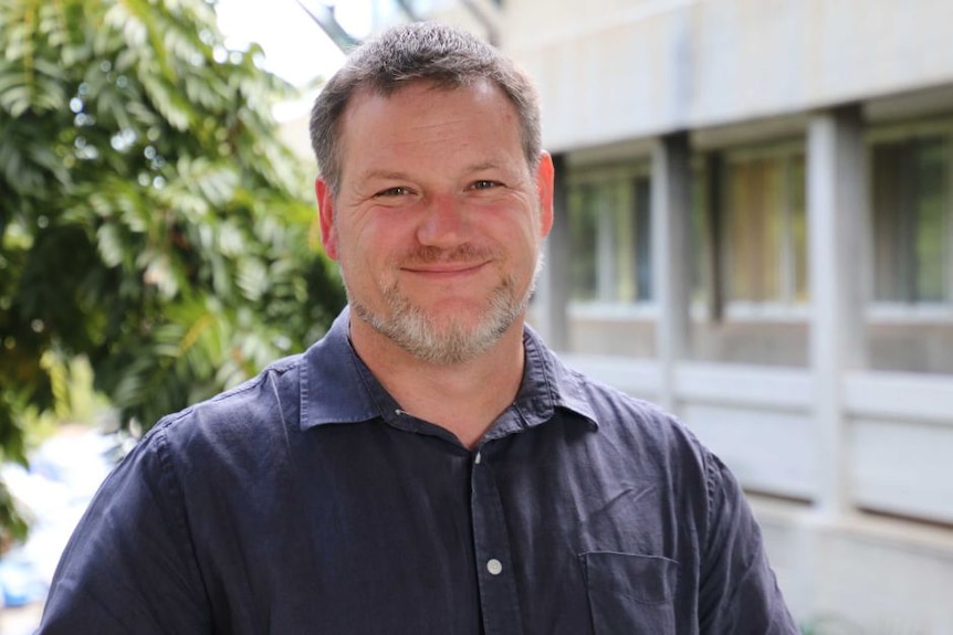 Portrait of a man in his forties with a grey beard and brown hair. He has a navy button up shirt on.