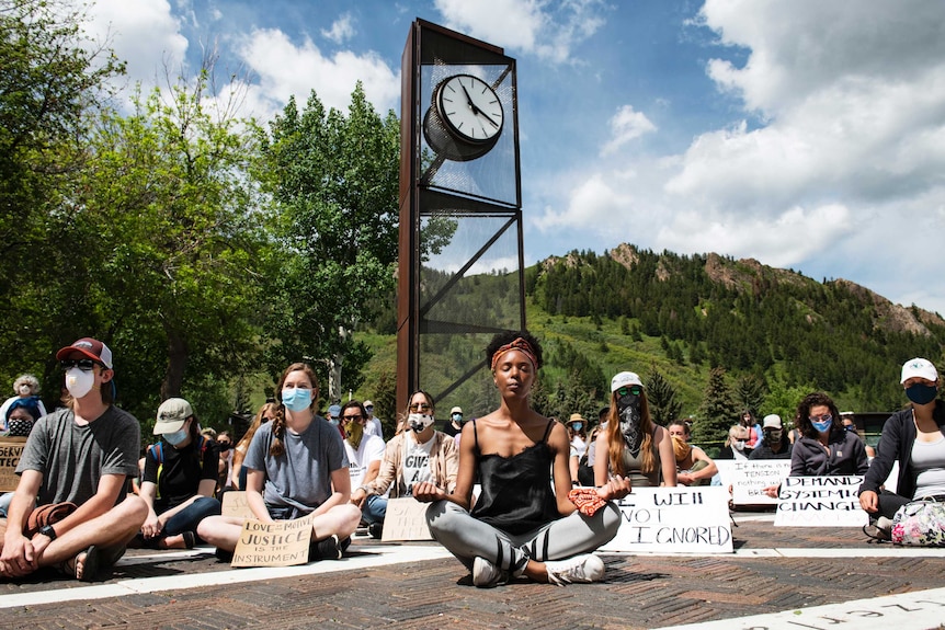 Dozens of cross-legged people meditate in front of a tree-covered hill, with protest signs in front of them.