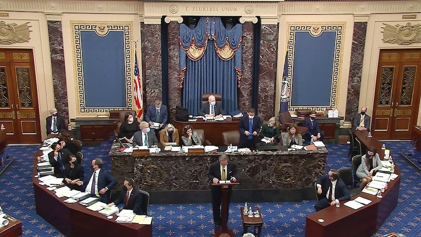 An aerial shot of a full senate chamber shows people sitting at benches as a man stands in the middle of the floor and speaks.