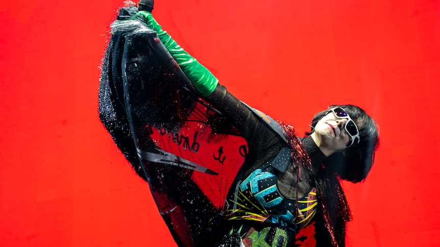 Yeah Yeah Yeahs' Karen O raises her hand, wearing a cape, DIY jumpsuit and shades with a red background live on stage