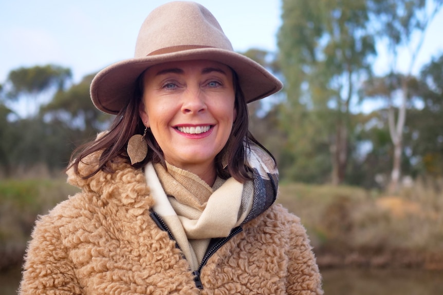 A woman in a felt hat and woolly jumper in the bush smiling into the camera.