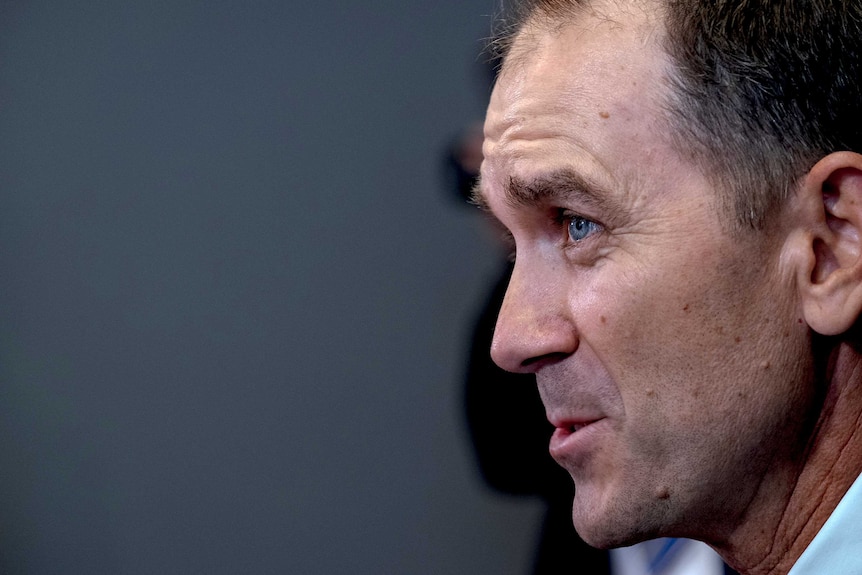 A side-on head shot of Justin Langer talking against a grey background.