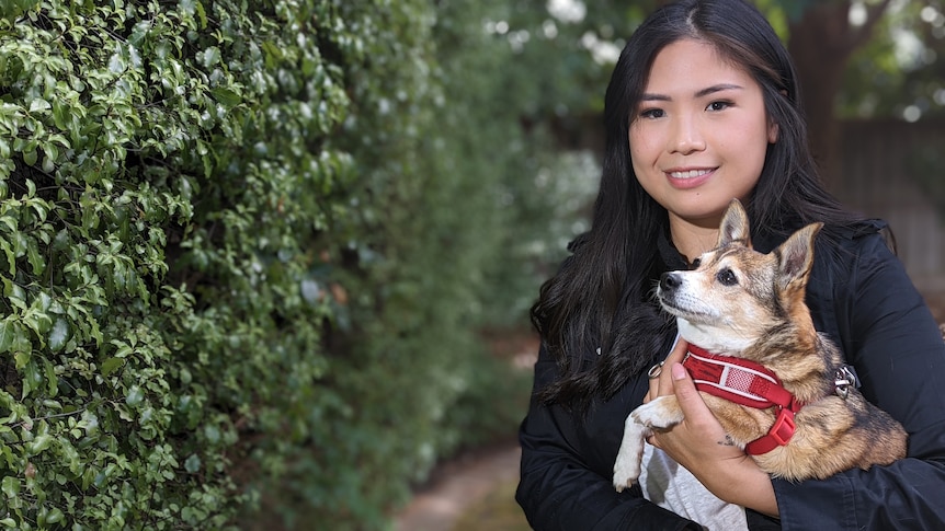 A smiling woman stands in a garden holding a small dog in her arms 