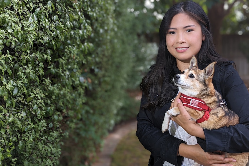 A smiling woman stands in a garden holding a small dog in her arms 