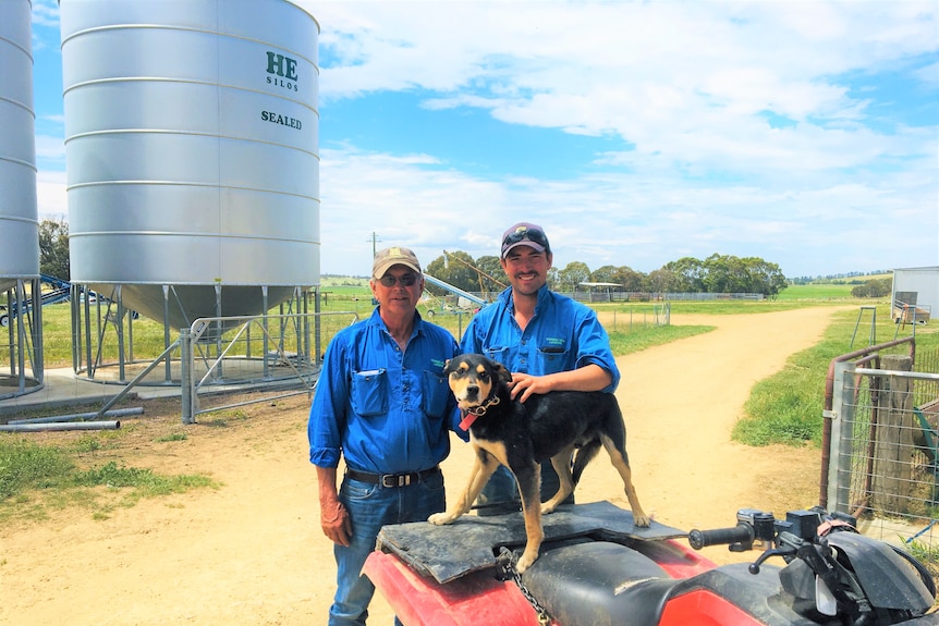 Two men in a blue shirts standing behind a quad bike with a black dog on top.