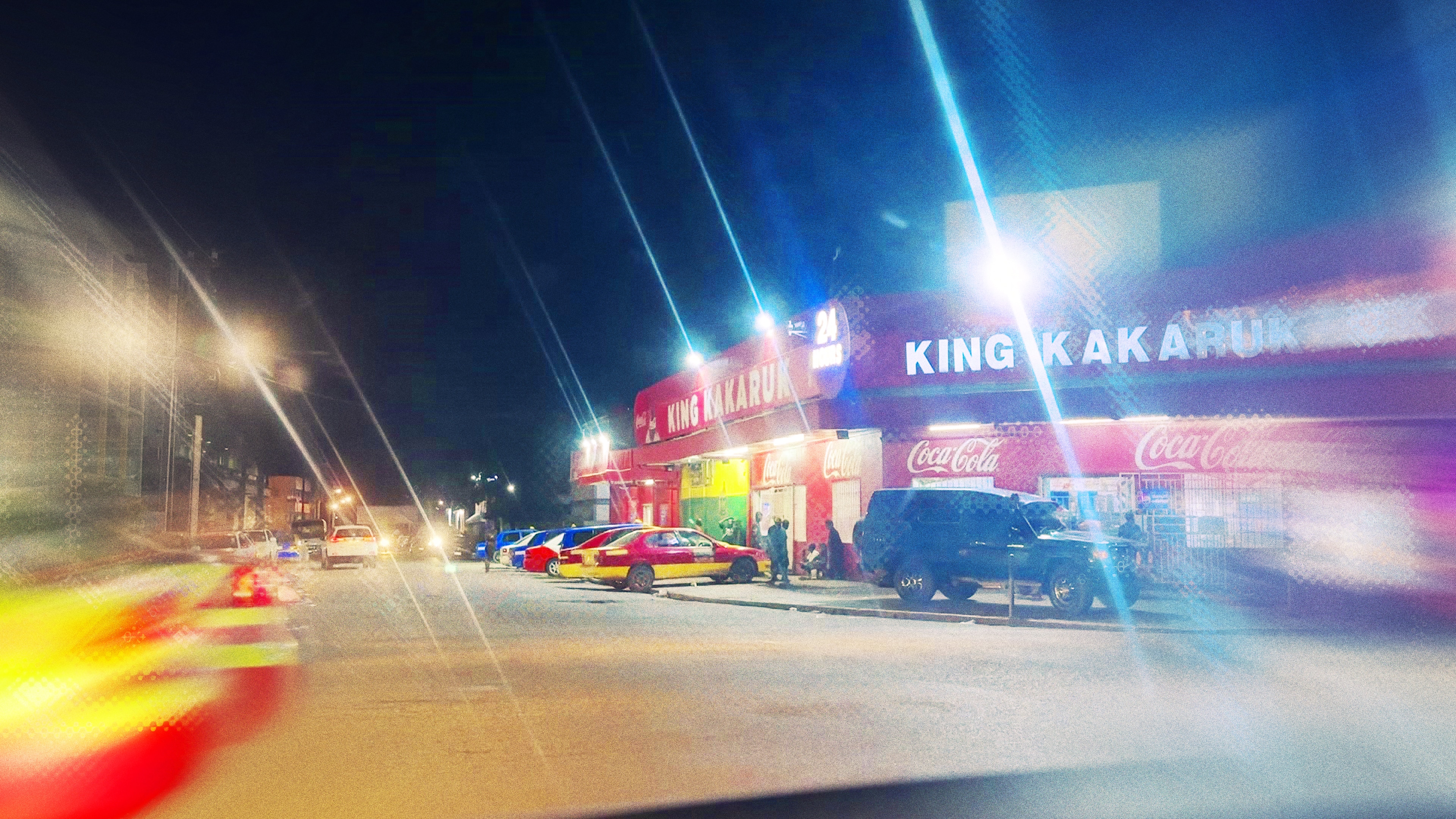 Por la noche, los coches se aparcan delante de una tienda roja y la gente se arremolina en el sendero cercano.