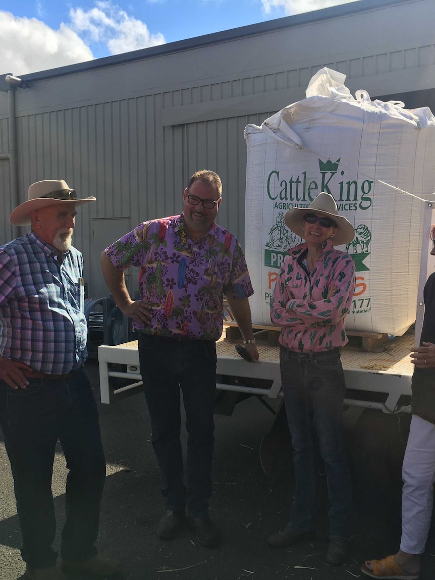 Seven farmers and Rotary members gather around the back of a truck with a large bag of feed on it, smiling and laughing.