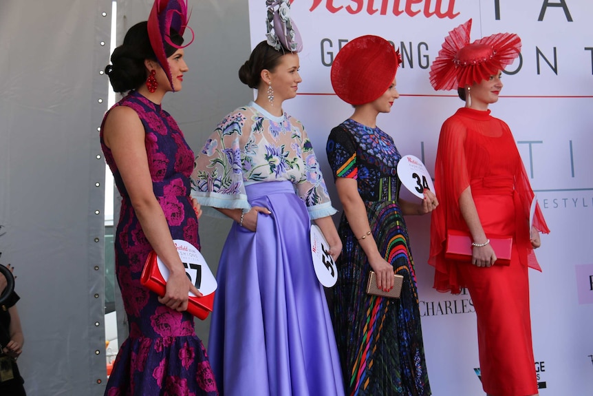 Ladies dressed in beautiful outfits stand on a stage, each holding a big number.