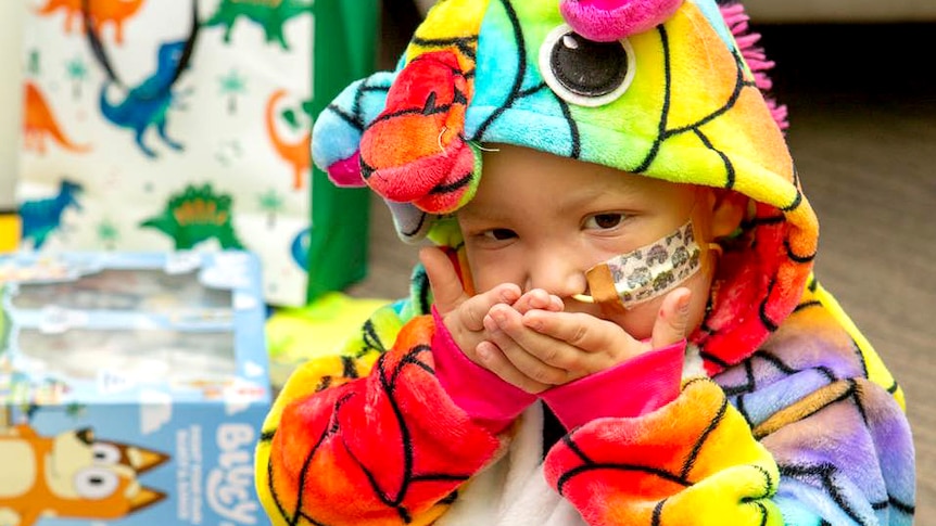 A child with an oxygen tube in his nose wears a onesie that looks like a rainbow unicorn.