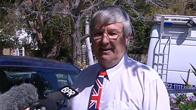 A grey-haired man in an Australian flag patterned tie stands in front of reporters' microphones on a suburban street.