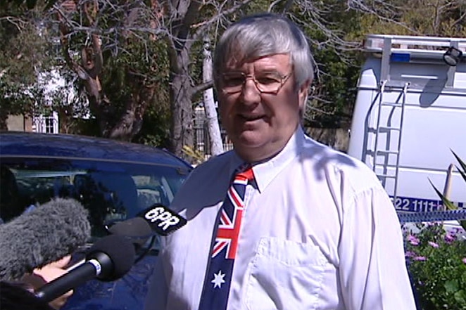 A grey-haired man in an Australian flag patterned tie stands in front of reporters' microphones on a suburban street.