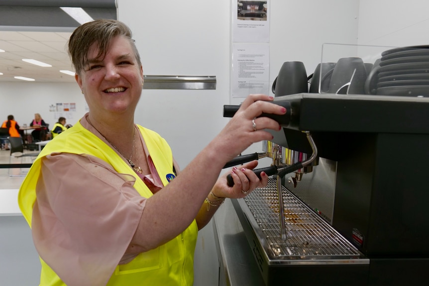 Kylie is at work making coffee, wearing a high visibility vest and smiling at the camera.