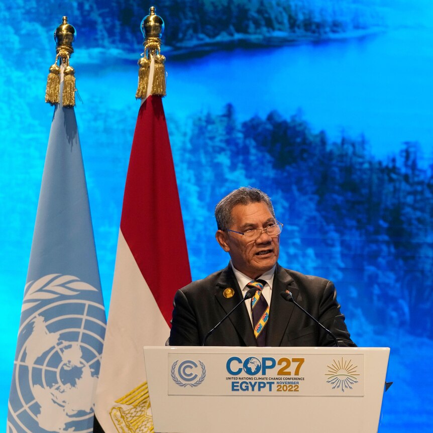 A man in a suit speak from behind a lecturn while standing before two limp flags.