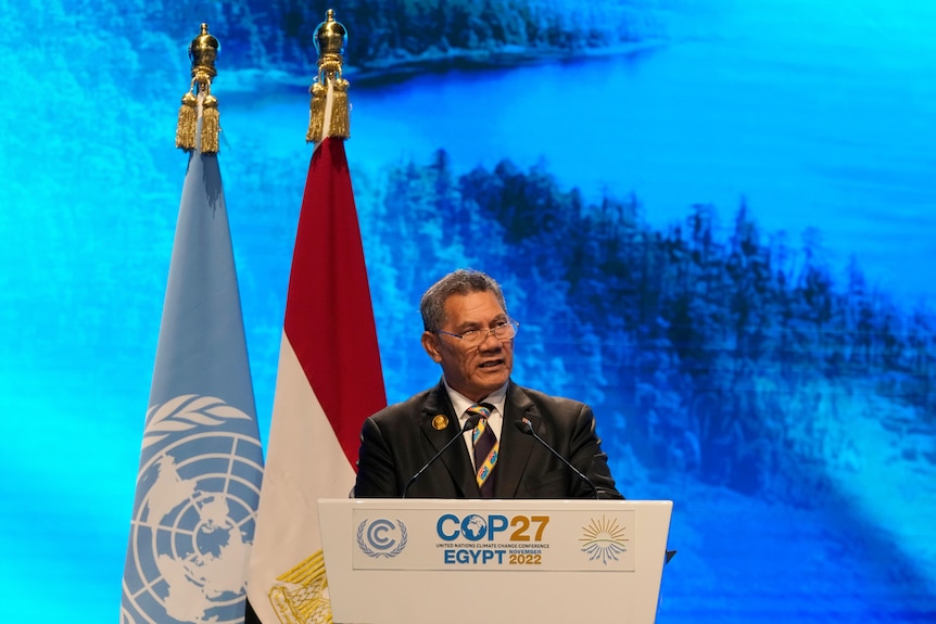 A man in a suit speak from behind a lecturn while standing before two limp flags.