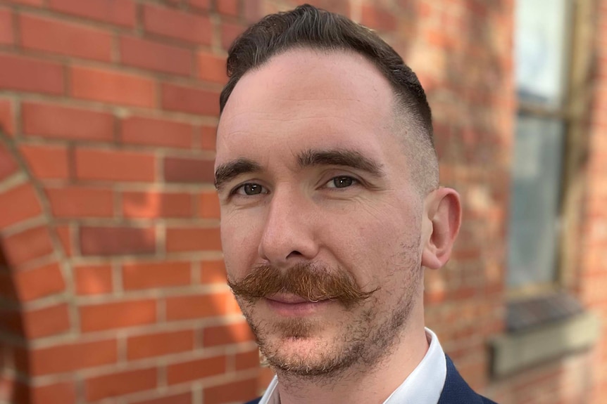 A man with a moustache wearing a blue blazer stands against a brick wall