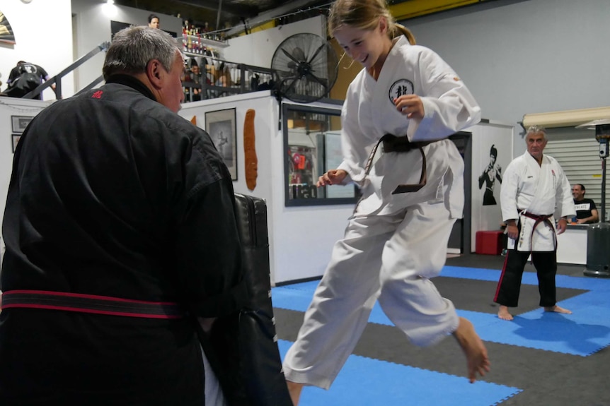 A girl martial arts students kicks a foam pad being held by an instructor.