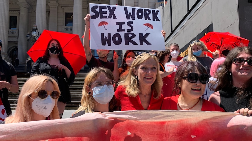 A group of women holding a large banner smile in the sun on the steps on parliament.