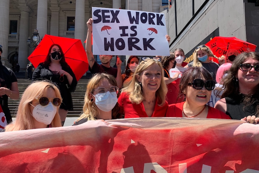 A group of women holding a large banner smile in the sun on the steps on parliament.