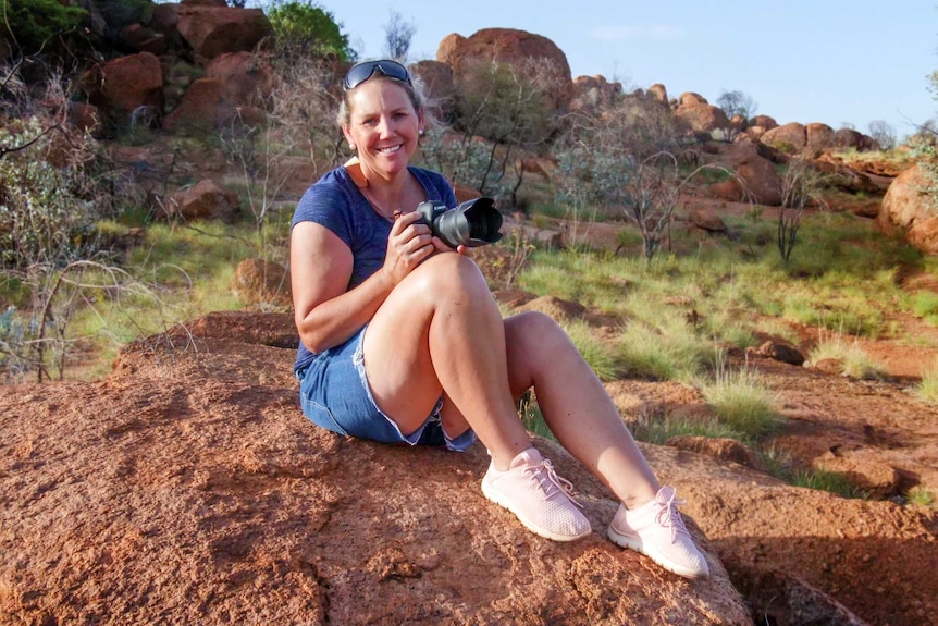 Mount Isa midwife Andrea Mitchell sits on a boulder in the outback holding a camera.
