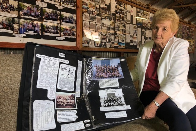 A woman in her late 70s sits holding a large black scrapbook about the history of a town called Yaappet. 