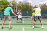 Four older people play ball on a tennis-style court using paddles similar to ping pong paddles