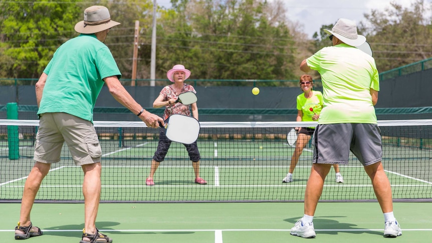 Four older people play ball on a tennis-style court using paddles similar to ping pong paddles
