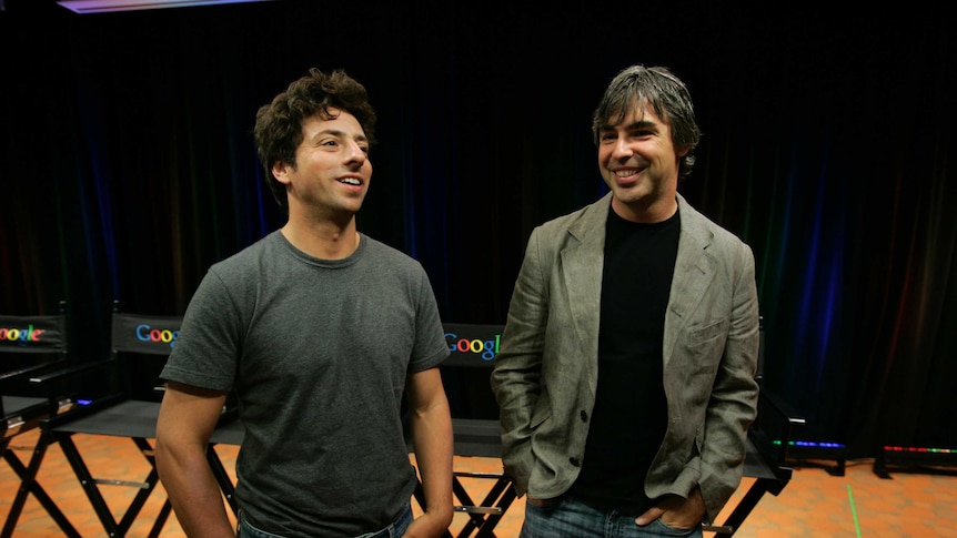 Two men stand on a stage in front of black director's chairs reading 'Google'