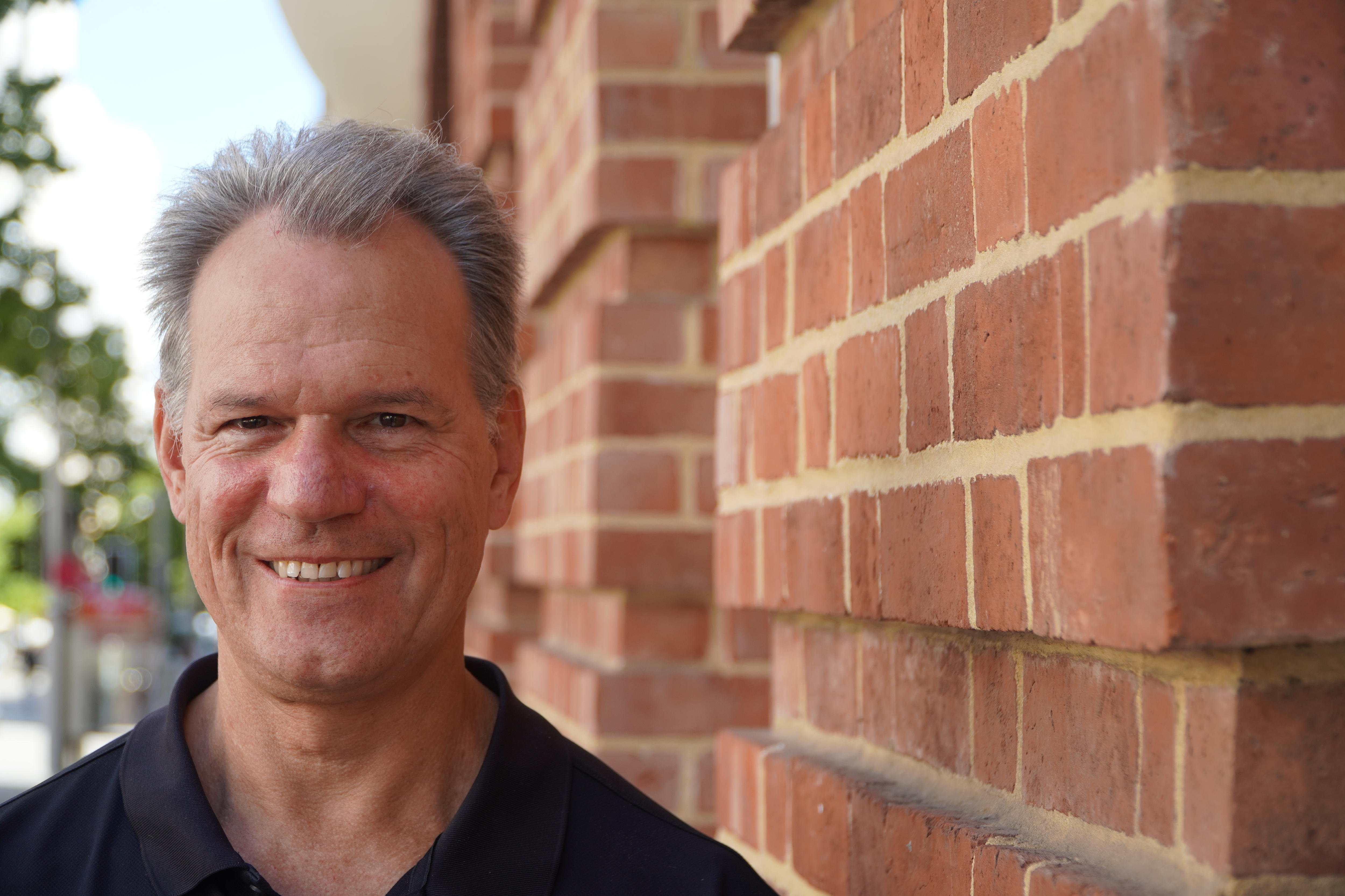 Eric Lilford smiles next to a brick wall. 