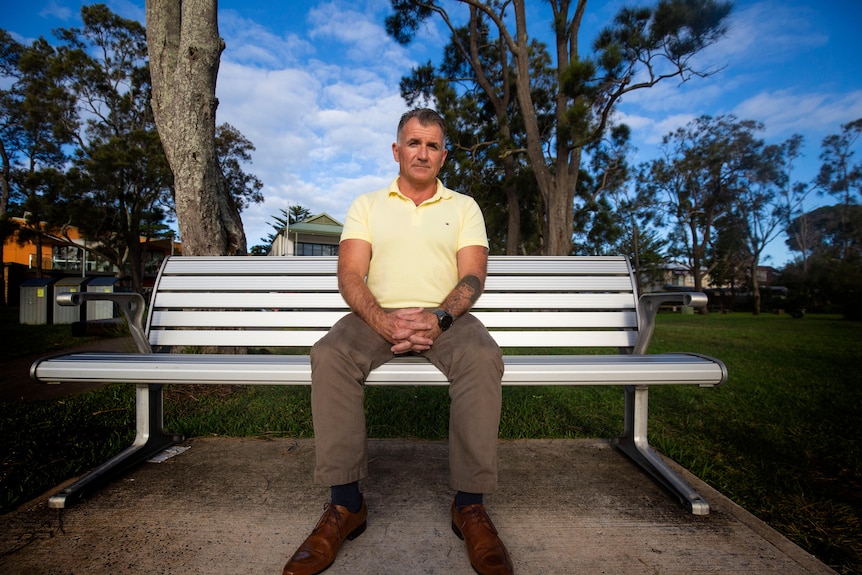 Lee Sarich, in grey pants and yellow shirt, sits on a silver park bench with hands folded over lap, and neutral expression.
