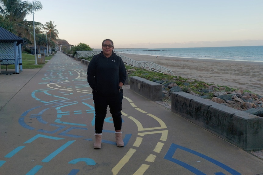 Sophie standing on a footpath next to Yeppoon main beach at sunset, ocean and horizon behind her.