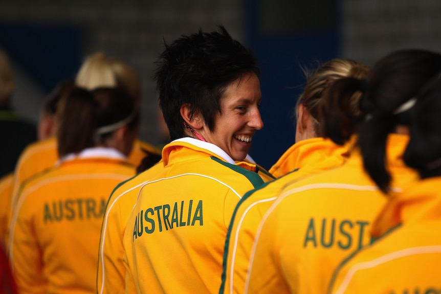 A woman with short black hair wearing a yellow and green Australia jacket smiles over her shoulder