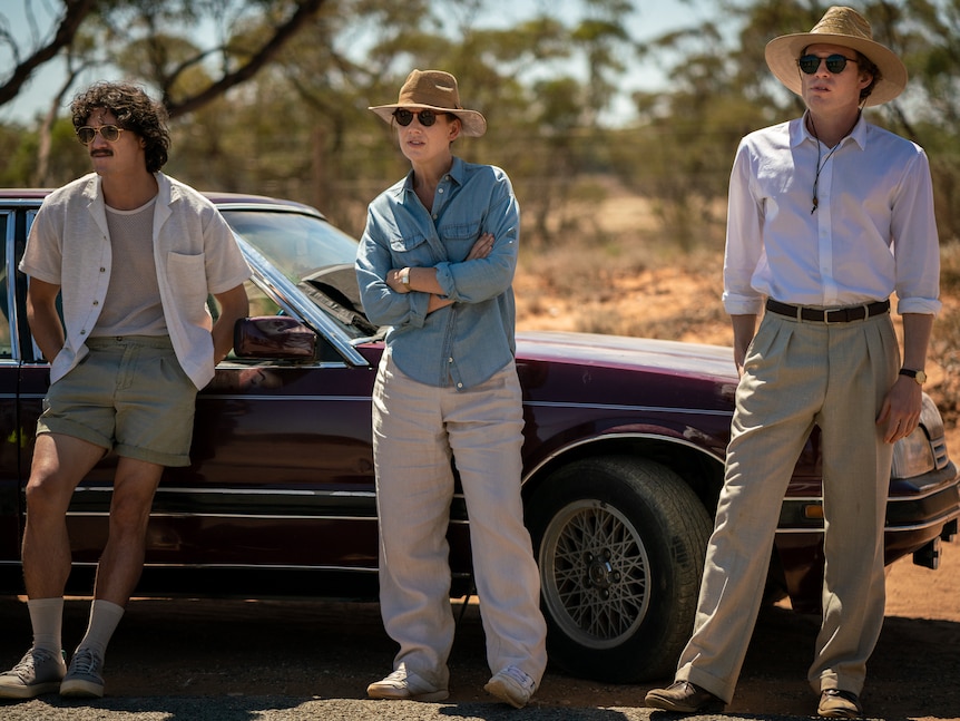 Three people stand alongside a car in hats and sunglasses, looking away into the distance