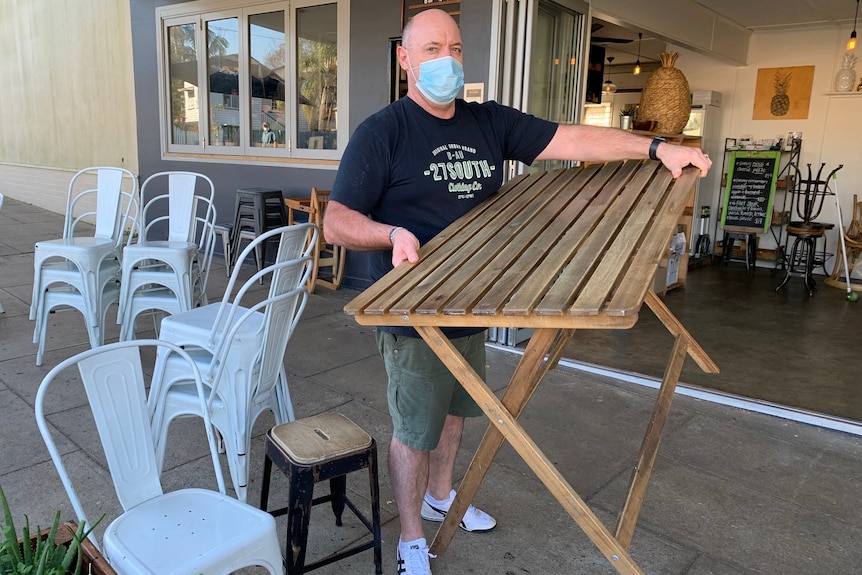 A man wearing a mask carries a wooden table at a cafe as he prepares to close