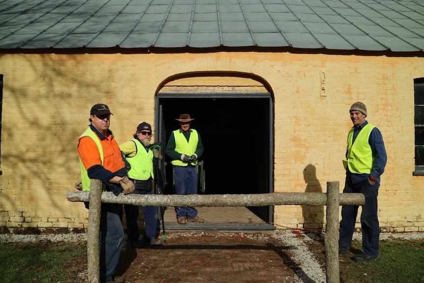 Four men wearing hi-vis vests stand outside a shearing shed