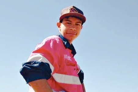 A young boy in a pink and blue shirt looks down and smiles at camera with blue sky above