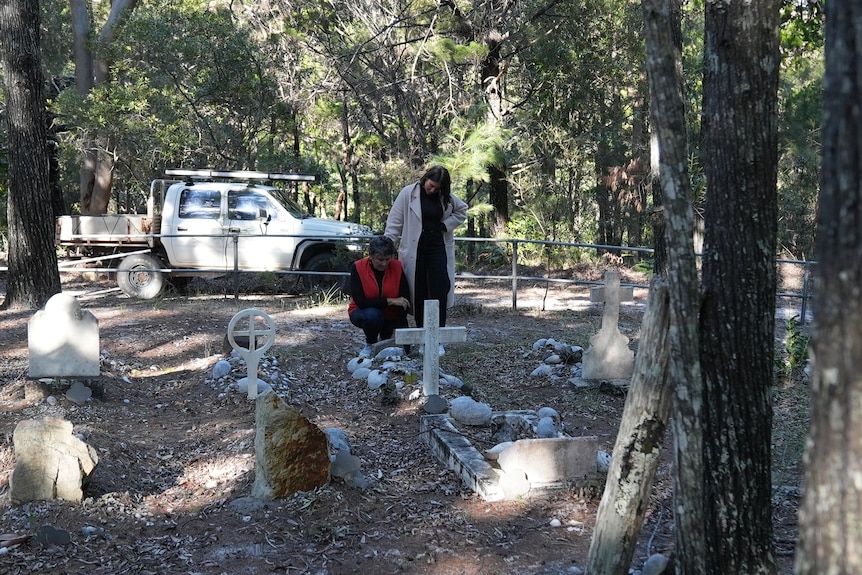 A 71-year-old woman kneels down to pay her respect to her grandmother while her adult daughter stands over her. 