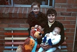 Mark James (l) standing on a colourful striped bench next to his mother, Maria, who has his brother, Adam (r), in her arms.