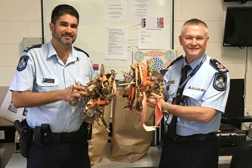 Two police officers hold handfuls of homemade slingshots surrendered to them by children from Yarrabah.