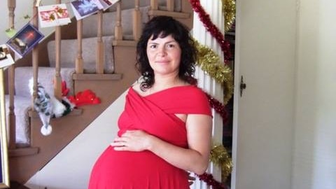 A heavily pregnant woman wearing a red dress stands in front of a staircase festooned with cards and tinsel.