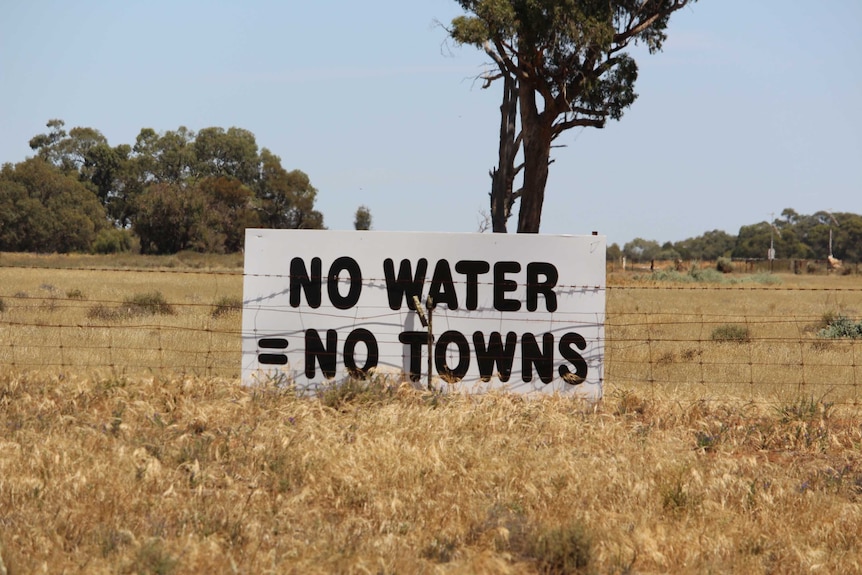 A sign that reads: No water = no towns sits on a fence on a dry irrigation paddock
