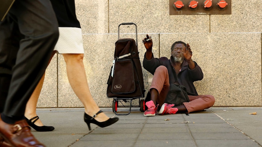 African-American sits on the pavement with a trolley waves to a man in a suit and a white woman wearing professional clothes