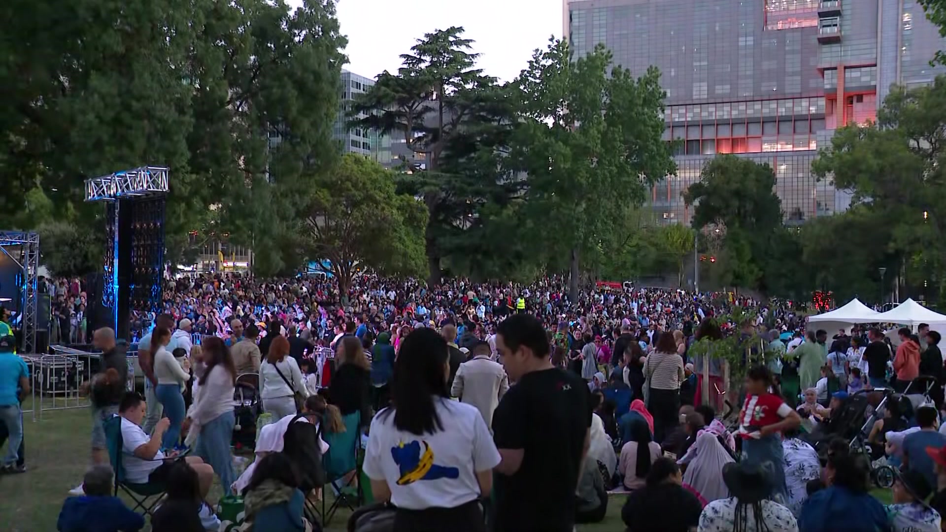 Grandes multidões cobrem a grama de um parque da cidade onde foram montados um palco e algumas pequenas tendas, com arranha-céus atrás.