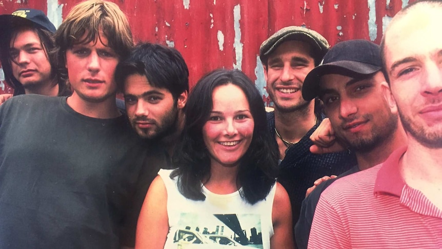 The Cat Empire and manager stand, smiling in front of a red corrugated iron shed