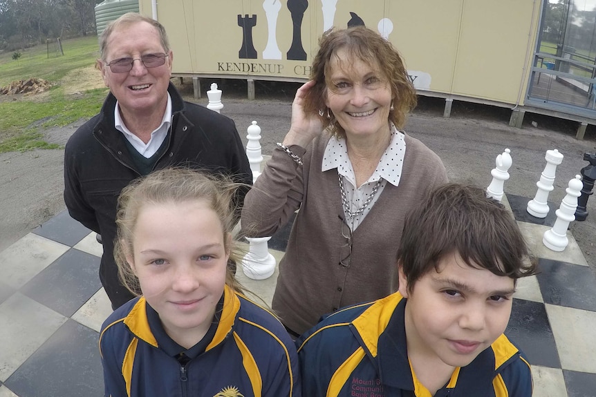 Two adults stand behind two children on a huge chess board.