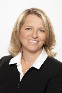 A headshot of a blonde woman wearing a black jacket with a white shirt.