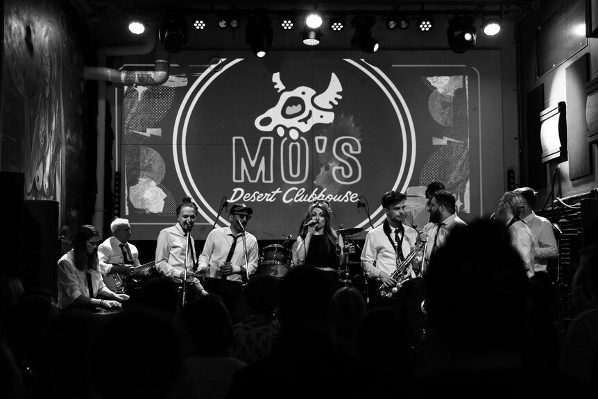 A band of eight musicians performing on stage in black and white in front of a sign that says mo's.